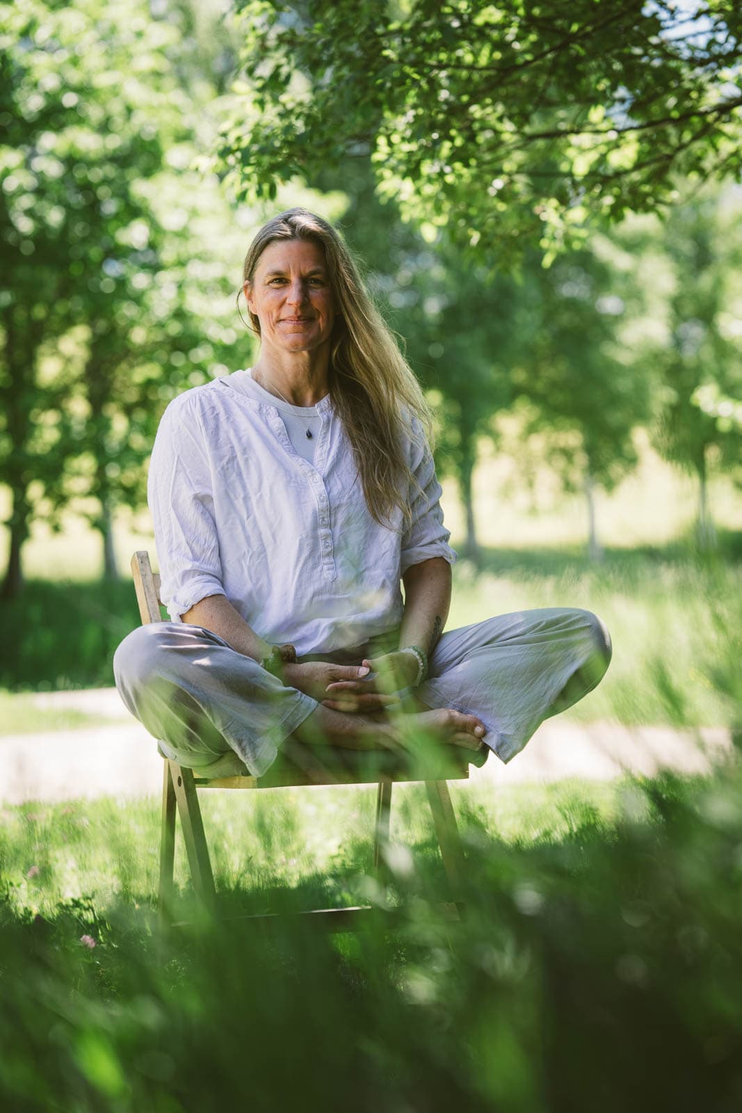 Caroline sitzt im Schneidersitz auf einem Holzstuhl im grünen Sommerlaub, warmes natürliches Lächeln.