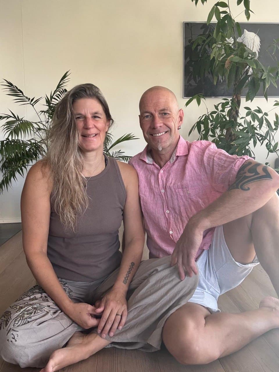 Caroline und Klaus sitzen nebeneinander auf dem Holzboden im Studio und lächeln in die Kamera.